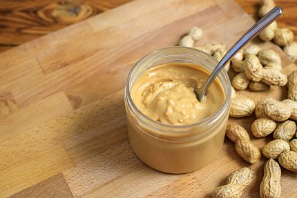 A glass jar of peanut butter with a spoon sticking out of it. The jar sits on a wooden board, with peanuts scattered to the right.