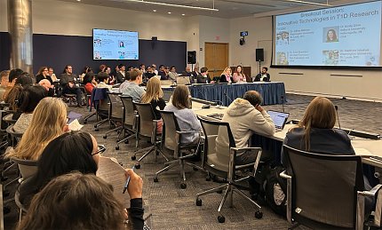 Attendants at a horseshoe-shaped table, with more rows of participants in chairs around the perimeter of the room. A projector screen is visible in the corner of the frame.