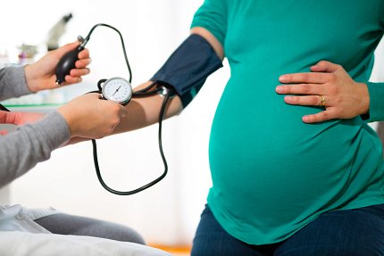 A health care worker (offscreen aside from their forearms and hands) measures the blood pressure of a pregnant patient. The patient is wearing a green shirt and holds one hand to their belly.