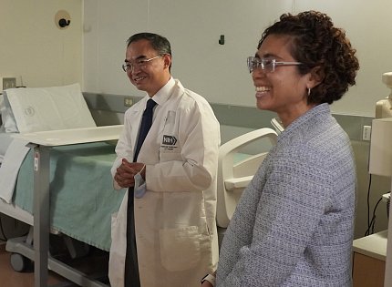Chung and a researcher in a white lab coat smile at someone out of the left frame. A hospital bed sits in the background.
