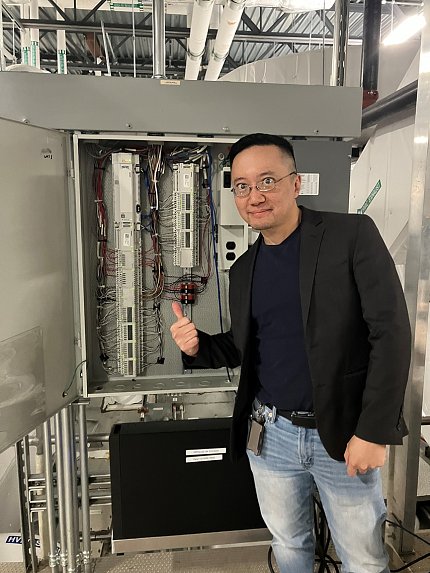 A man in bespectacled man in jeans and a black blazer gives a thumbs up to an open electrical panel behind him.