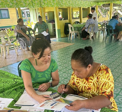 Choy, l, works with a woman completing a workbook. Other people are seated around the room with their own workbooks.