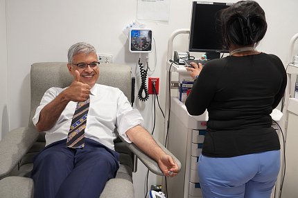 A man in a white shirt and striped tie seated in a room with medical equipment rolls up his shirt sleeves. A person partially out of frame with their back to the camera types on a computer.