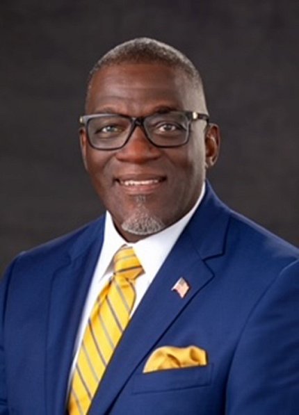 Headshot of Clark, wearing glasses and a navy suit with a yellow, striped tie.