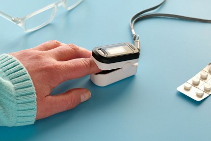 A person's left hand rests on a light blue surface. The tip of the pointer finger is inside the pulse oximeter device.