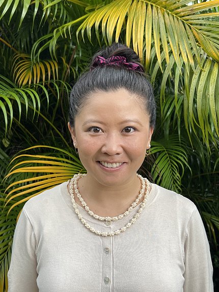 A smiling Choy poses in front of green and yellow palm fronds. She wears a tan cardigan and a double-stranded seashell necklace.
