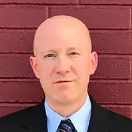 Headshot of Hartzell, who is wearing a suit and tie and standing against a brick wall.