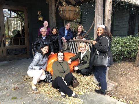 A group of 9 individuals arranged around a large orange pumpkin. The pumpkin rests on a bed of straw, to the right of a wooden bench. A line of bushes and a dark green building fill the background. 