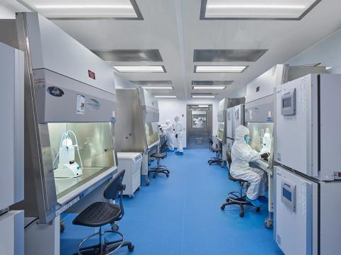 Scientists in white PPE suits and masks sit and stand in front of fume hoods in a brightly lit room.