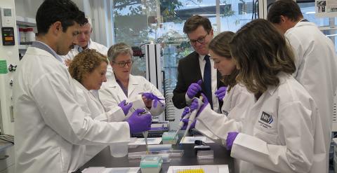 The group stand around a table in white lab coats using pipettes.