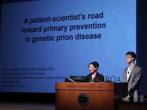 Vallabh and Minikel stand behind a large lectern. Behind them is a large projector screen featuring the title of their talk.