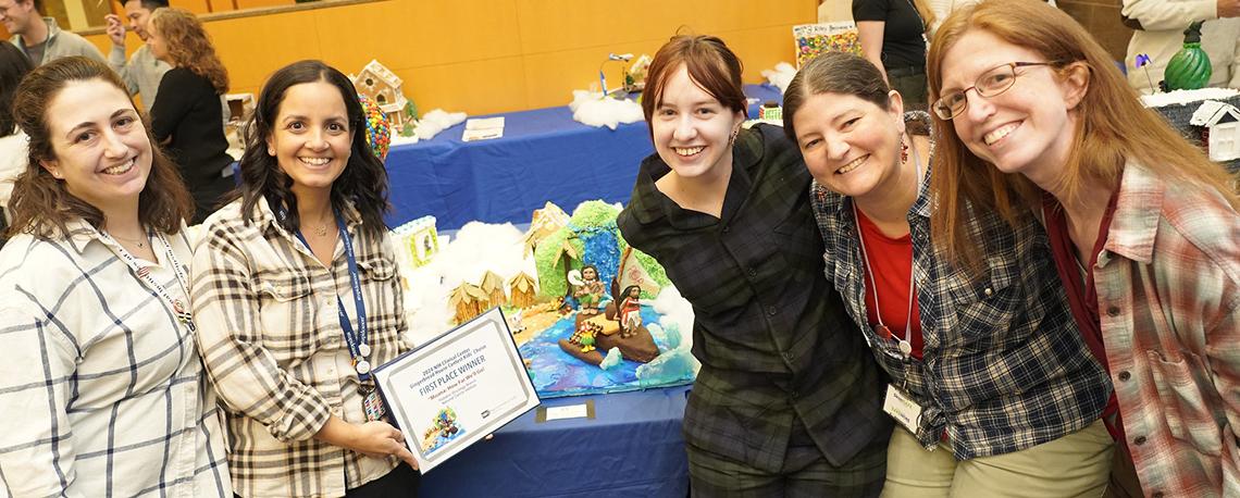 Five women smile next to their winning gingerbread; one holds their certificate.