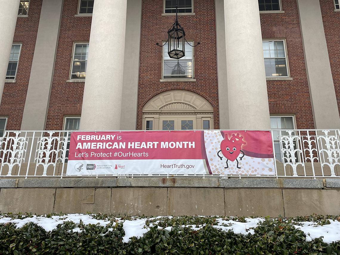 A red and white banner reading "February is American Heart Month" hangs from a white railing on the front of a brick building with large white columns.