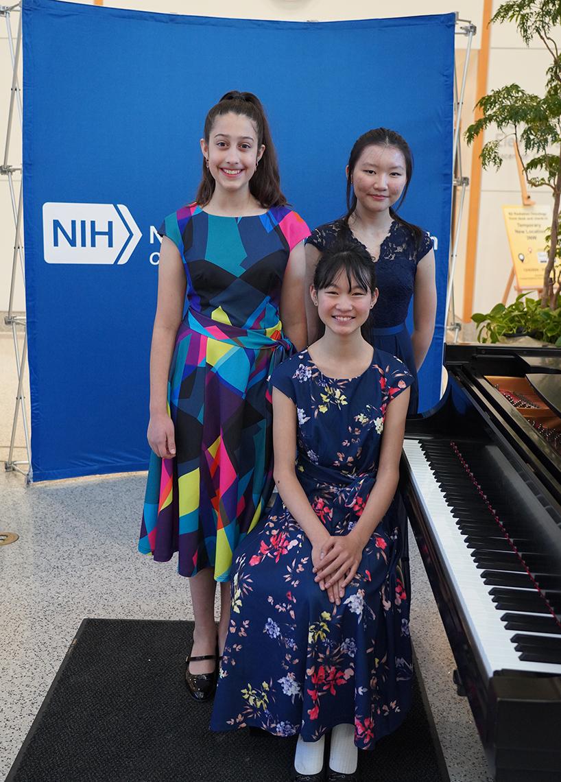 Three young, smiling pianists next to piano in atrium