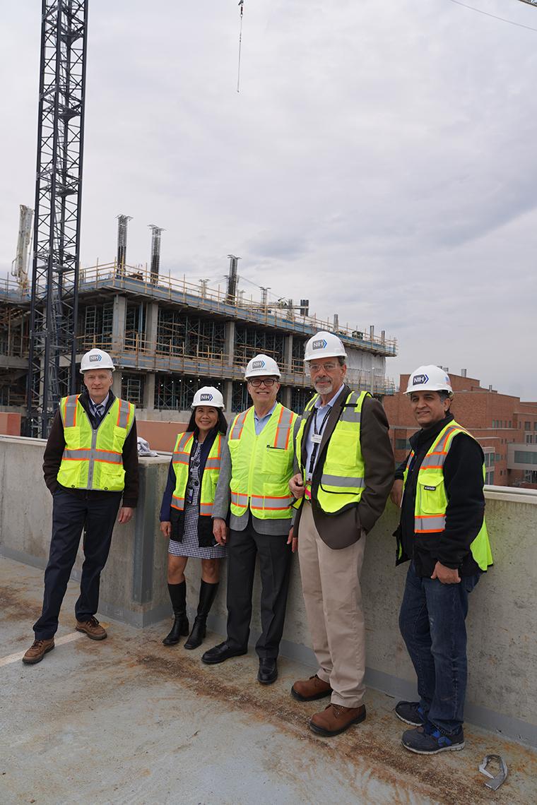 Five people in white hard hats and fluorescent yellow construction vests stand against a concrete barrier. The frame of a partially built, multi-story building rises behind them.