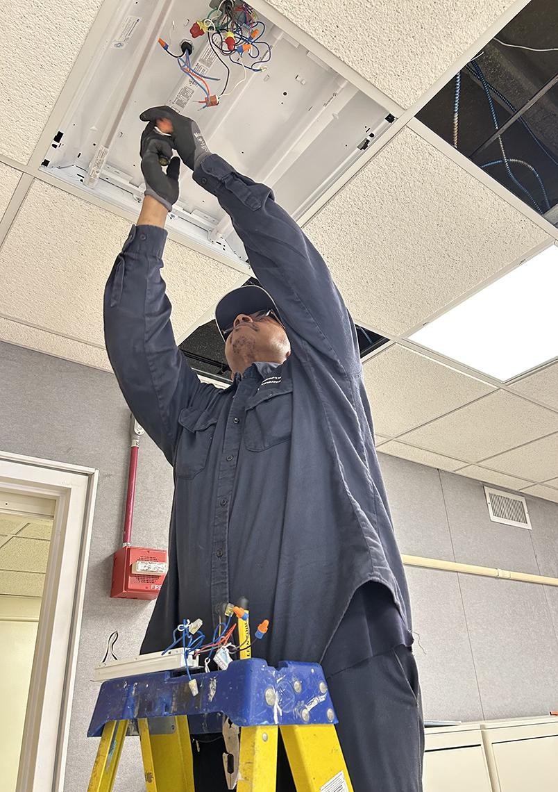Hoes stands on ladder in an NIH office holding tools up to wires in ceiling.