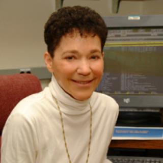 Nussinov seated in a red chair; she is wearing a cream-colored turtleneck and a long chain necklace.