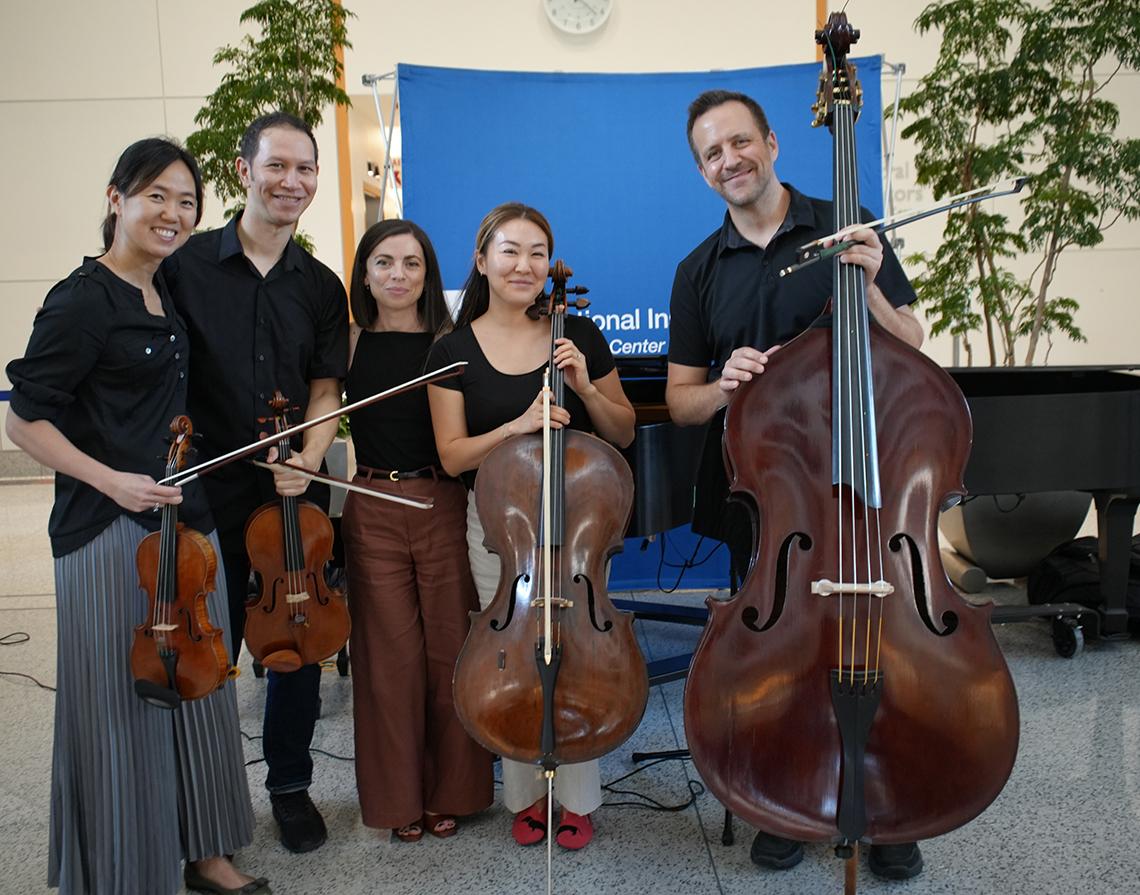 Five musicians stand with their stringed instruments in the Clinical Center atrium.