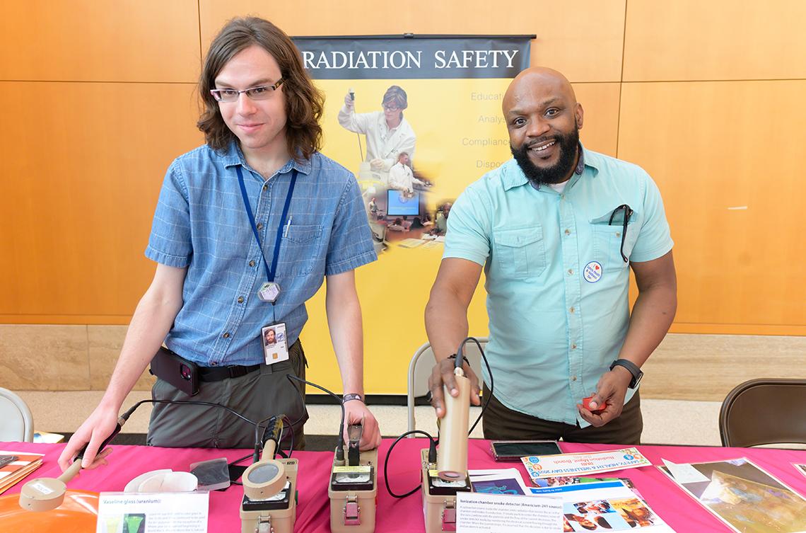 Two colleagues hold meters, tools with Radiation Safety poster behind.
