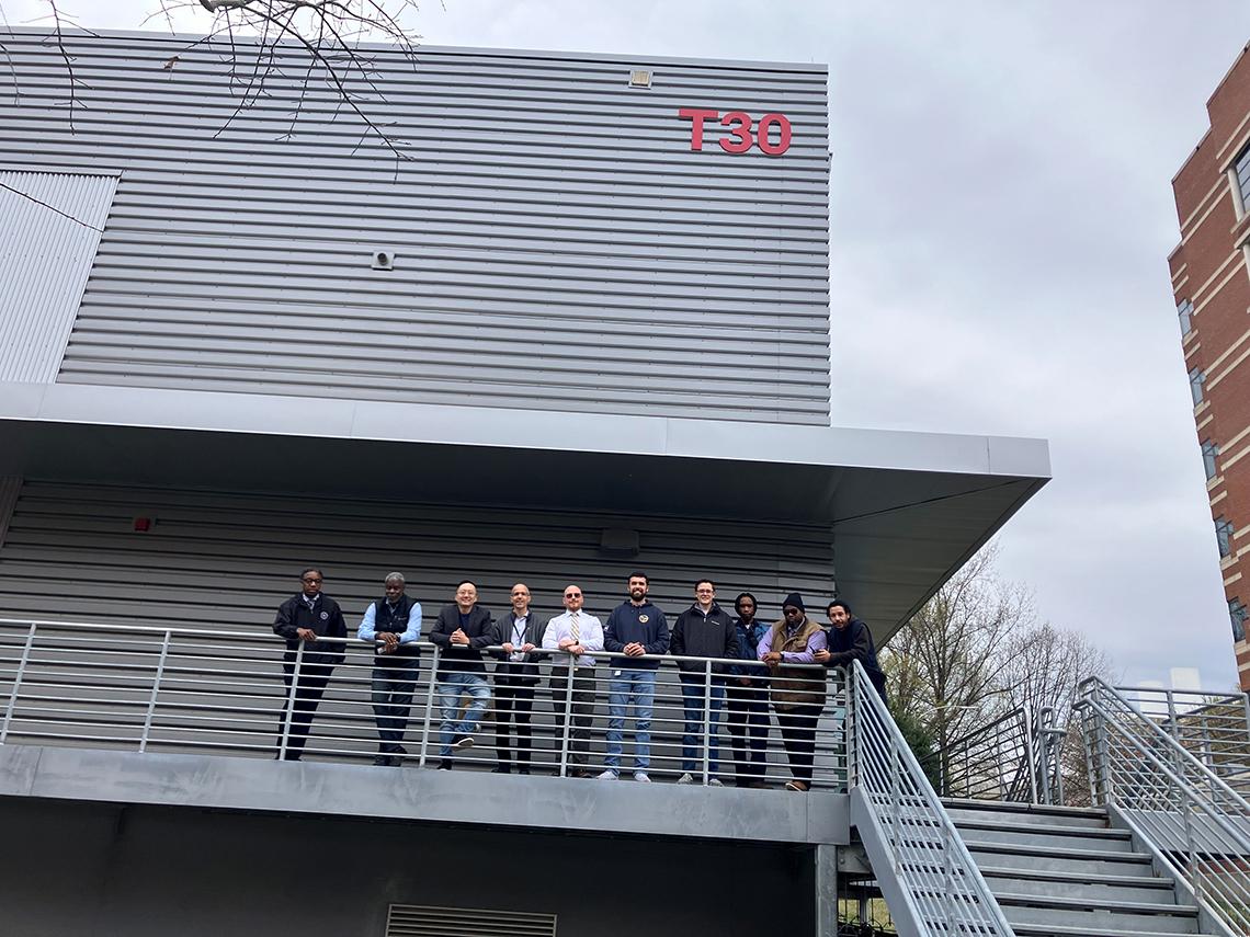 A group of 10 men lean against the railing of a walkway along the outside of a metal building. The building number, T30, is shown in red letters in the upper right-hand corner near the roof.