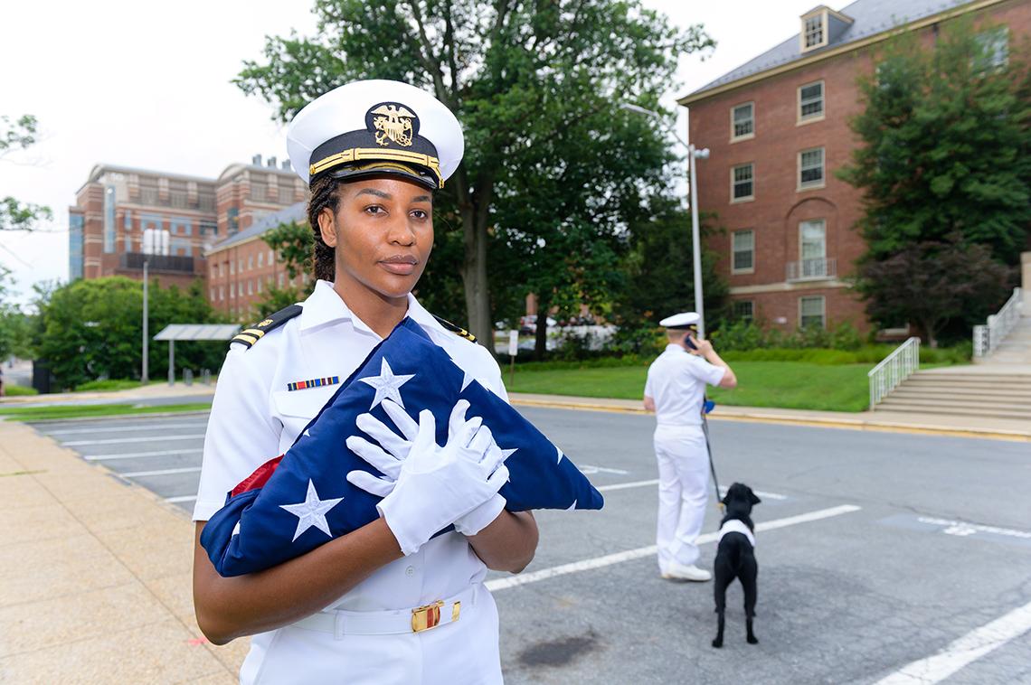 A PHS officer holds a folded American flag. Another officer stands in the background with a black dog.