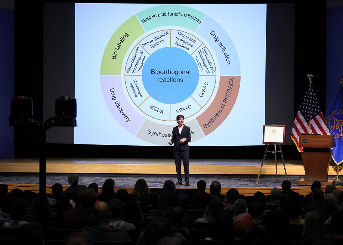 Bertozzi stands on a stage silhouetted by her PowerPoint presentation, which features a diagram of different bioorthogonal reactions.