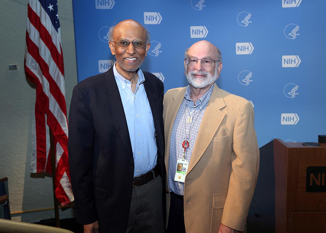 Goldstein and Nath stand in front of a light blue backdrop patterned with the NIH and HHS logos.