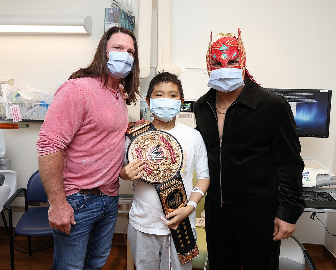 Two wrestlers pose with child, holding a title belt, in the patient's hospital room.