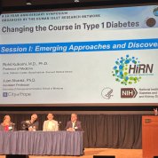 A speaker at a podium on the right side of the photo gestures as he presents; a table of four listens from the left side of the image. A projector slide above them shows the presenter's slide show.