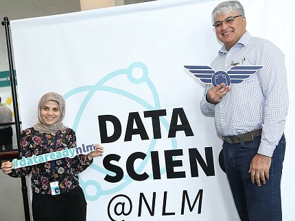Dr. Ghada Zamzmi Alzamzmi and Dr. Sameer Antani of NLM Two attendees stand in front of a sign that reads "Data Science @ NLM"