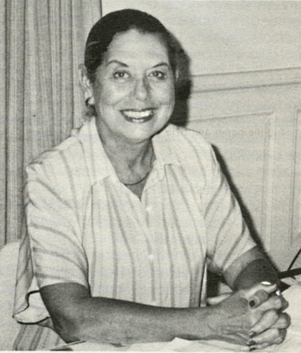 Belia “Bel” Ceja Black & white photo of a smiling Bel Ceja, sitting hands clasped at her desk